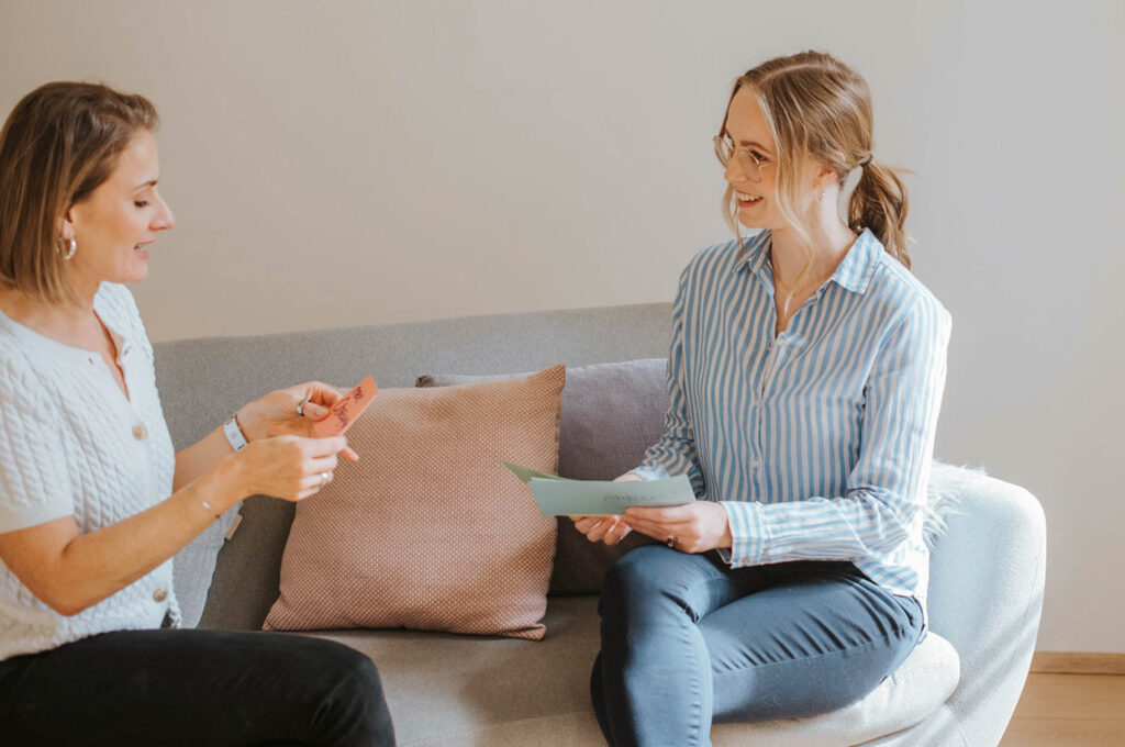 Die psychosoziale Beraterin Florentina sitzt einer Klientin gegenüber auf einer Couch. Beide Frauen haben die Beine überkreuzt, sind zueinander gedreht und sind in einem Gespräch. Dabei halten beide Karteikarten in den Händen. Die Beraterin lächelt ihre Kundin ermutigend an.