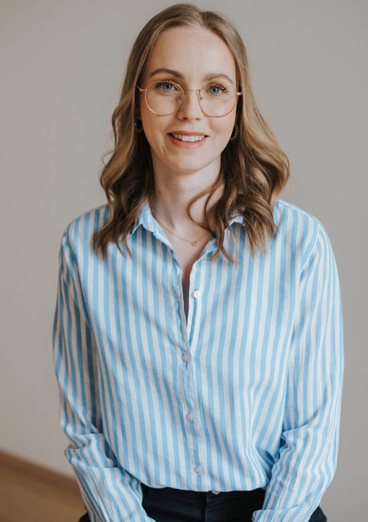 Die Gesundheits und psychosoziale Beraterin Florentina trägt eine moderne Brille und eine blau gestreifte Bluse. Sie lächelt in die Kamera und wirk sympathisch. Sie steht vor einem weißen Hintergrund und das Foto geht bis zu ihrer Hüfte.