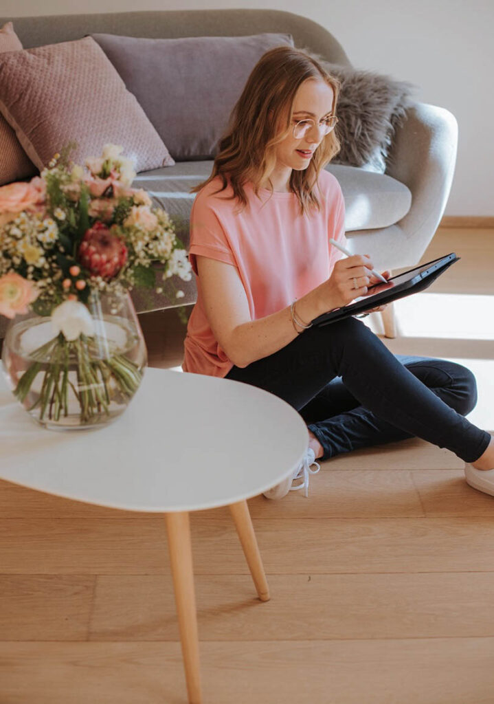 Beraterin sitzt vor einer couch auf dem Boden und arbeitet an ihrem iPad, im Vordergrund steht ein kleiner Tisch mit Schnittblumen. Die Trau trägt eine rosafarbene Bluse und eine dunkle Jeans.