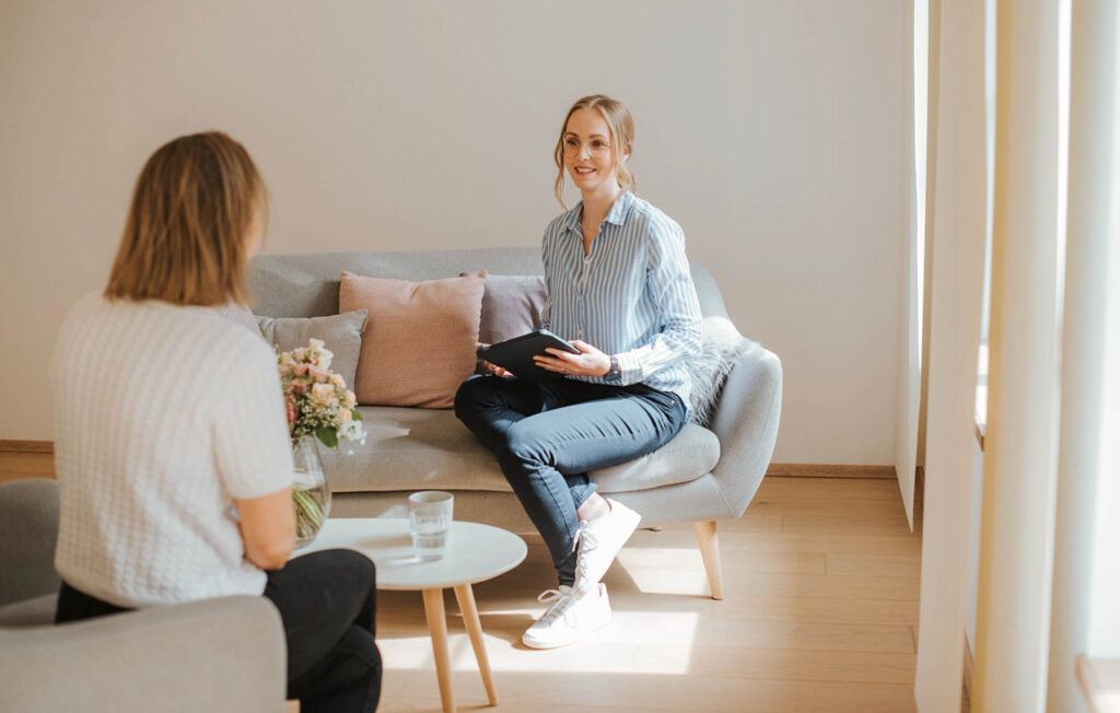 Die Beraterin Florentina für Gesundheitseinrichtungen sitzt einer Frau gegenüber. Zwischen den beiden Frauen steht ein kleiner weißer Tisch mit einer Vase Schnittblumen. Die Beraterin hält ein iPad in Ihrer Hand. Die Kundin ist ihr zugewandt.