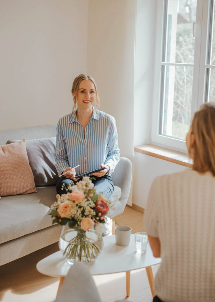 psychosoziale Beraterin Florentina hält ein iPad in ihrer Hand und sitzt auf einem Sofa. Ihre Kundin sitzt ihr gegenüber und ist mit dem Rücken zur Kamera gewandt. Zwischen den beiden Frauen steht ein kleiner Tisch mit einer Vase mit Blumen und einem Glas Wasser.