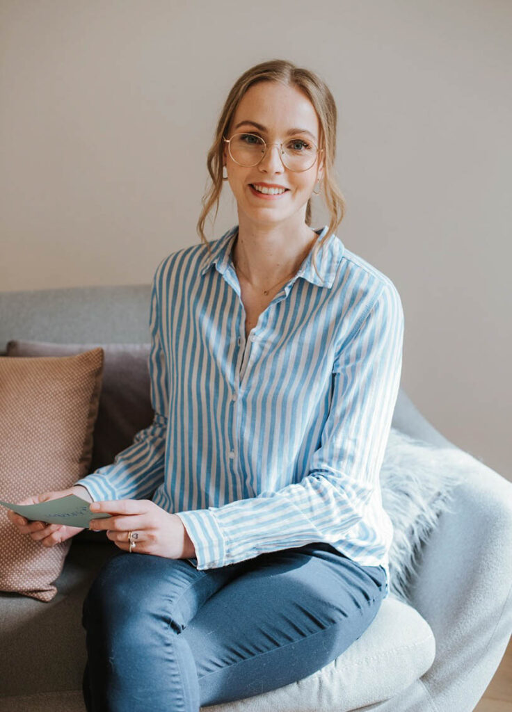 Psychologische Beraterin sitzt auf grauem Sofa, trägt Blaue und weiß gestreifte Bluse und hält eine Karteikarte in beiden Händen. Die Frau lächelt sympathisch in die Kamera und trägt eine moderne Brille.