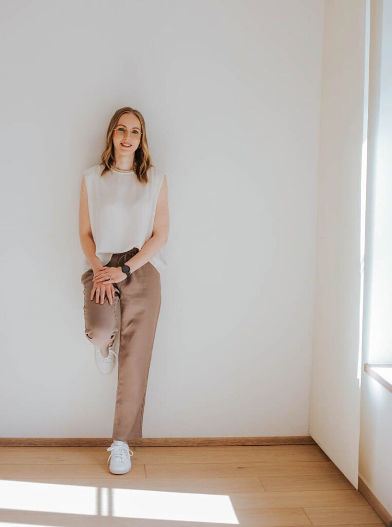 Die Beraterin Florentina steht vor einer weißen Wand und lehnt leicht an der Wand. Dabei hat sie ein Bein nach hinten an die Wand gestützt, das Andere ist ausgestreckt. Neben ihr ist der Anschnitt eines Fensters zu sehen, welches einen leichten Schatten wirft. Die Beraterin trägt eine weiße Bluse und eine braune Hose und lächelt in die Kamera.