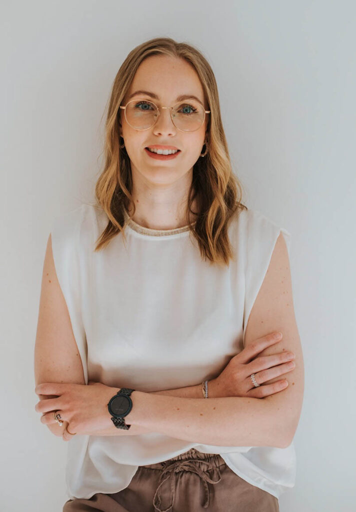 Die Beraterin Florentina lehnt an einer weißen Wand, hat ihre Arme leicht verschränkt und lächelt in die Kamera. Sie trägt eine weiße Bluse und eine braune Hose. Die Beraterin wirkt sympathisch und nahbar.