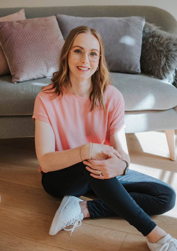 Die Beraterin Florentina für Gesundheitseinrichtungen sitzt am Boden vor einer couch und hat die Beine überkreuzt. Sie lächelt in die Kamera und trägt eine rosafarbene Bluse. Der Boden ist ein Parkettboden und es scheint ein leichter Schatten und Sonnenstrahlen auf sie.