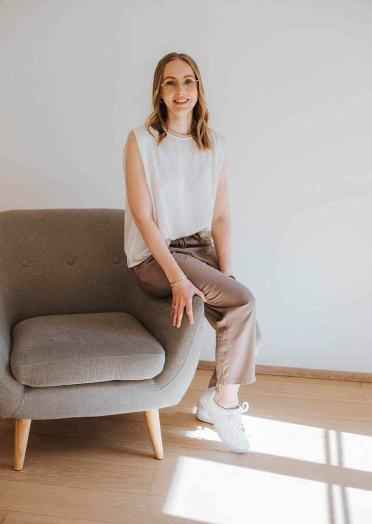 Die Beraterin Florentina sitzt auf einer Lehne eines braunen Stuhles und lächelt sympathisch in die Kamera. Sie trägt eine weiße Bluse und eine beige Hose und weiße Sneaker.