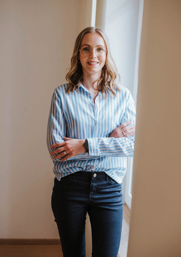 Psychologische Beraterin und Beraterin im Gesundheitswesen Florentina steht mit verschränkten Armen vor einem Fenster und sieht direkt in die Kamera, lächelt dabei leicht.